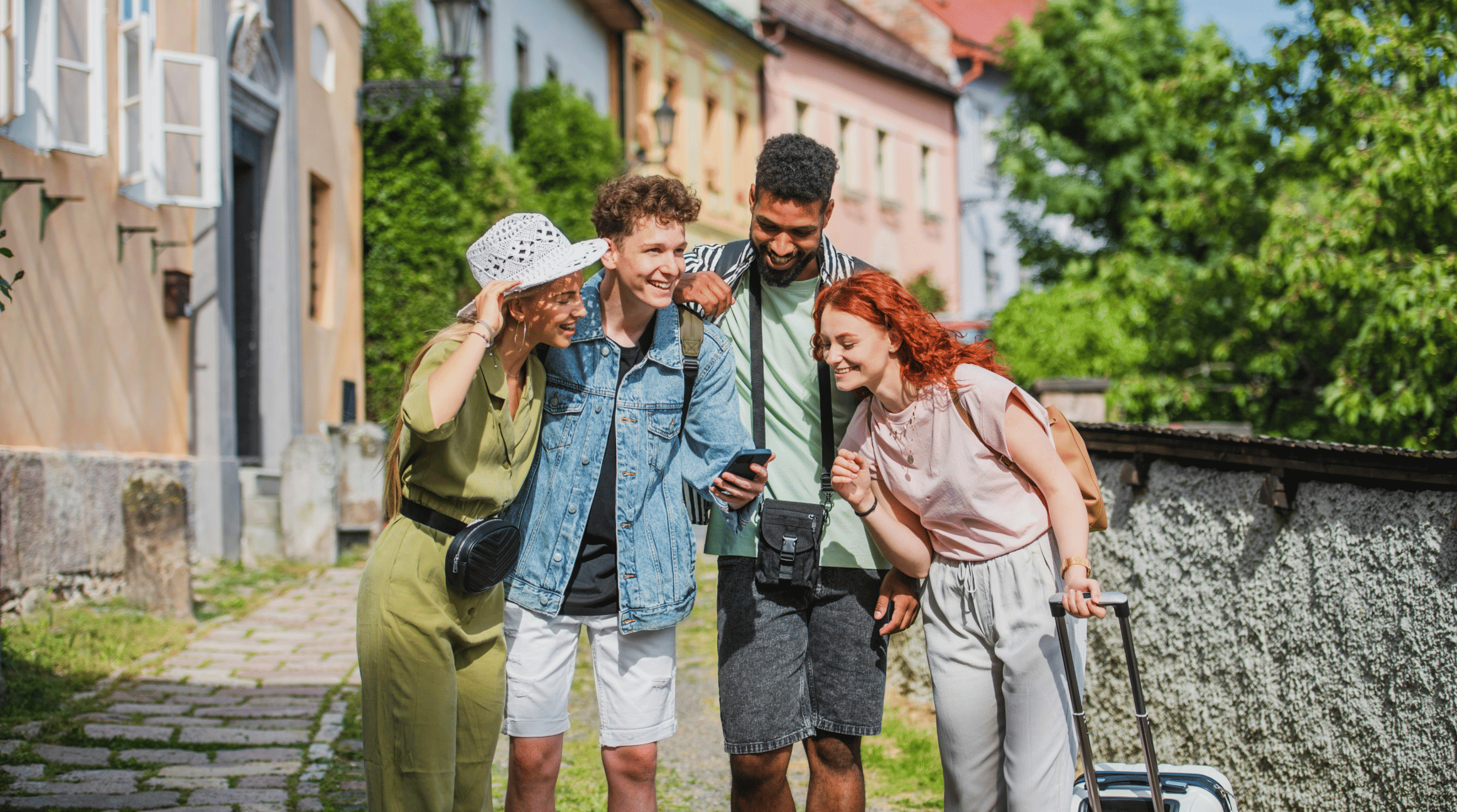 A colour photo of a group of four friends in the foreground. they are all dressed in casual clothes, in a group, and are looking at one mobile phone screen and smiling. In the background is a small, cute holiday village. To illustrate a blog post entitled 'The best group travel apps for 2025.'