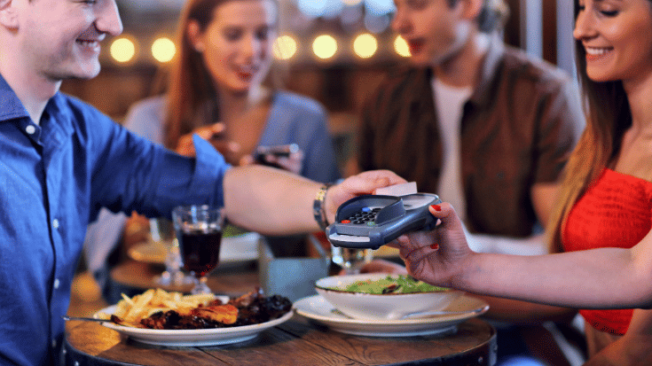 A colour photograph showing several friends sitting around a table in a restaurant laughing and smiling. The server's hand is visible in the foreground as she holds out a card machine. One of the diners is tapping his card to the machine, to pay for his share of the bill. To illustrate a blog post entitled 'The best group travel apps in 2025.'