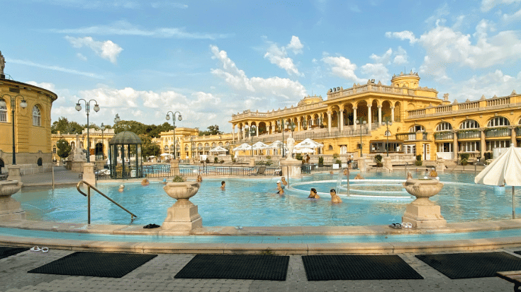 A photograph os the outdoor section of Budapest's Szechenyi baths on a sunny day. In the foreground you can see the bright blue waters of the thermal baths, with just four people walking and soaking in the water. In the backgroudn is the very distinctive and ornate yellow building of the thermal baths. To illustrate a blog post entitled 'The Ultimate Sziget Festival Guide.'