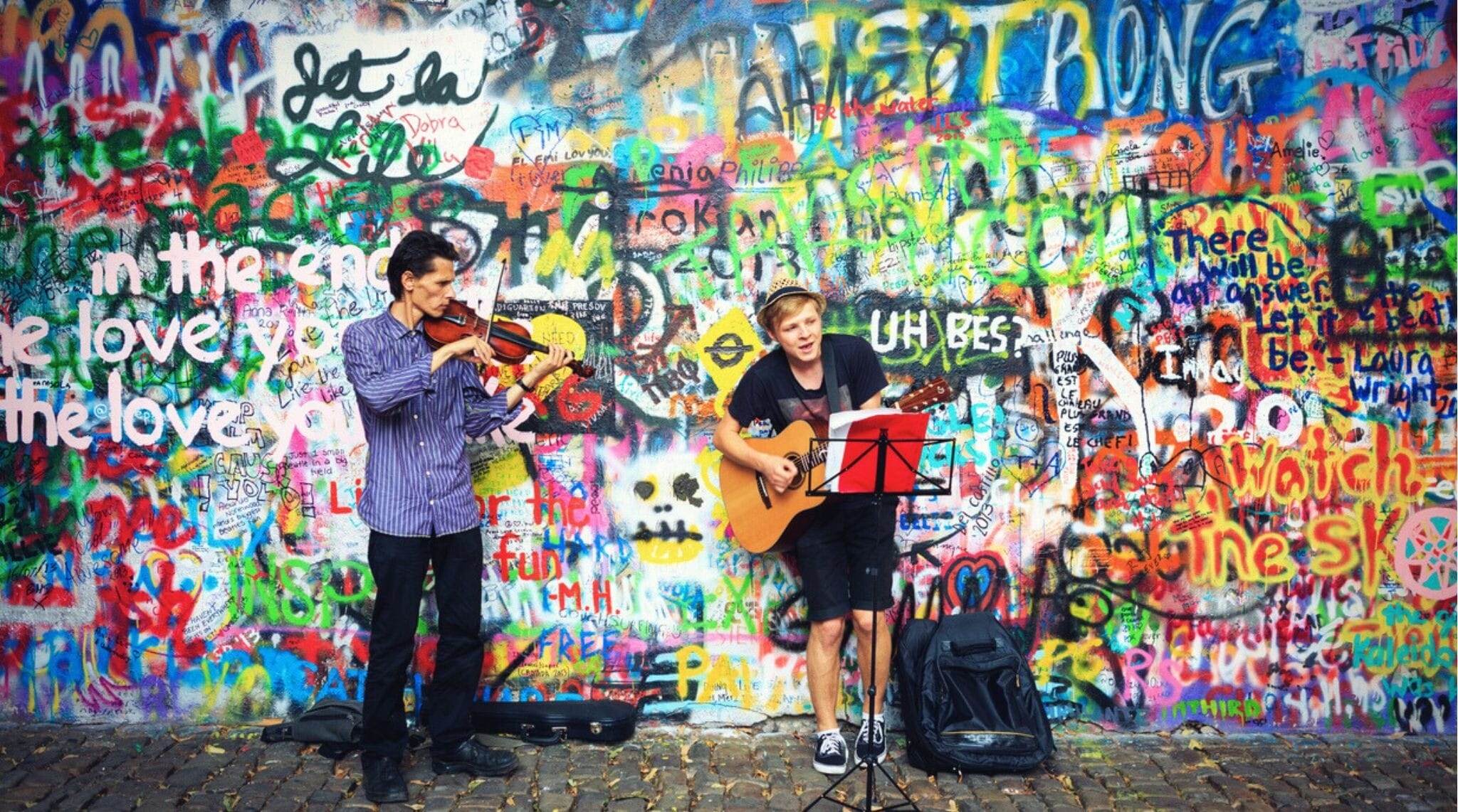 Dois jovens músicos tocando no Muro John Lennon, coberto por grafites inspirados em John Lennon e trechos de músicas dos Beatles.