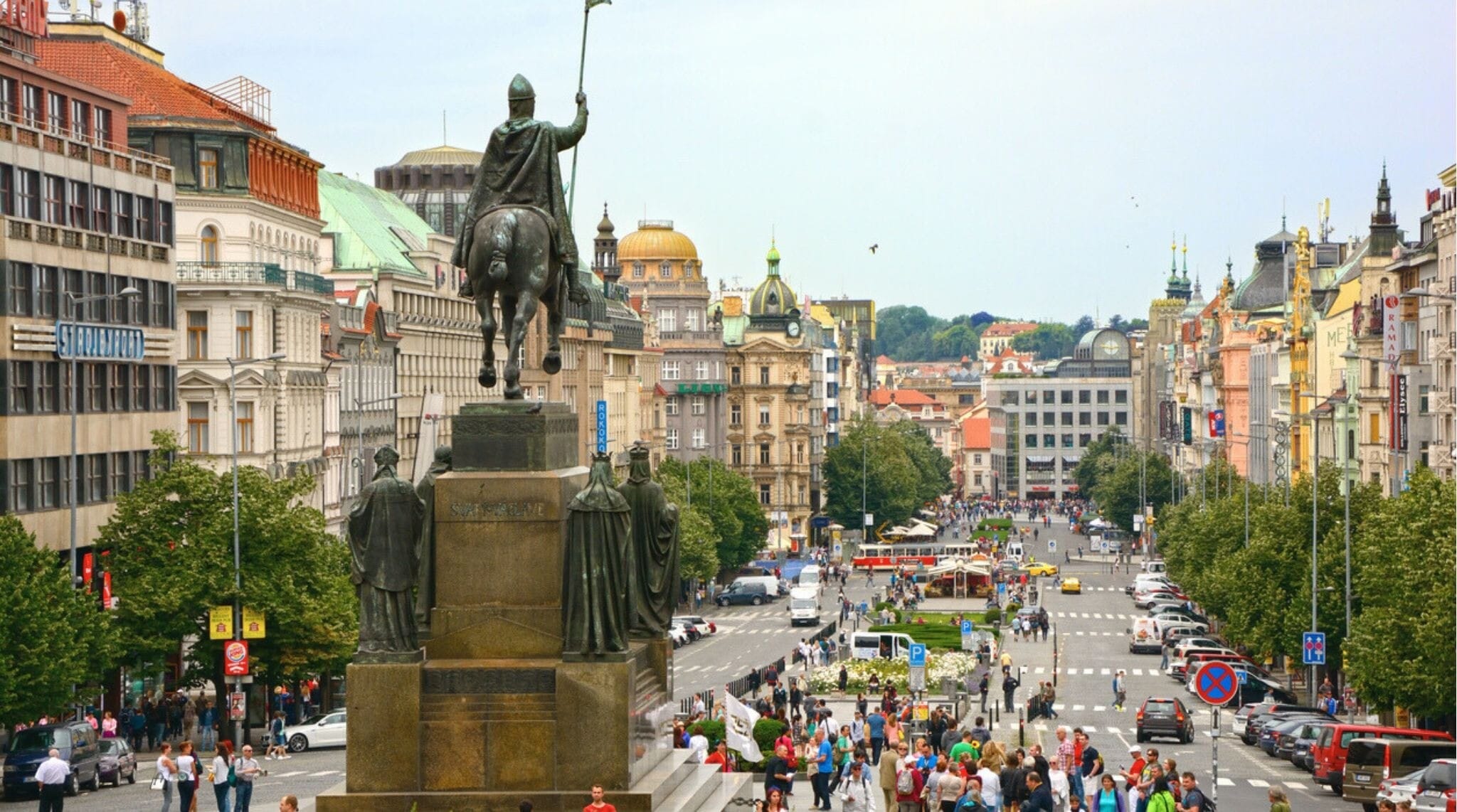 Praça Venceslau (Václavské Náměstí) — a principal avenida da cidade — vista a partir do Museu Nacional. A estátua de Venceslau aparece em primeiro plano à esquerda. Muitas pessoas caminham pela rua. Imagem horizontal.